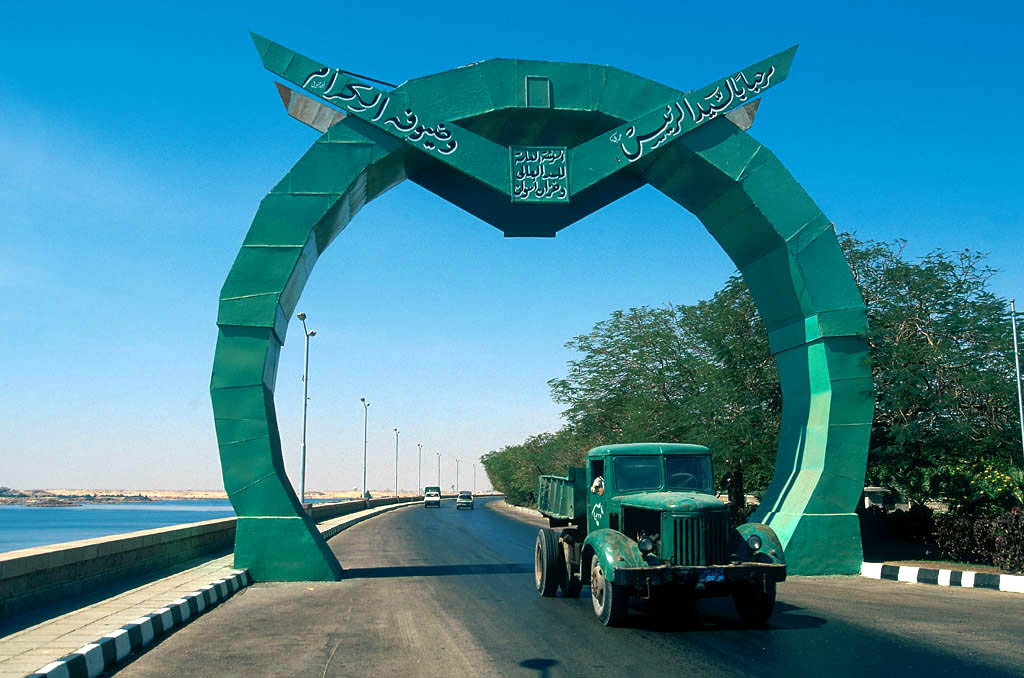 Une arche verte ornée d’inscriptions arabes enjambe la route du haut barrage d’Assouan, où circule un camion.